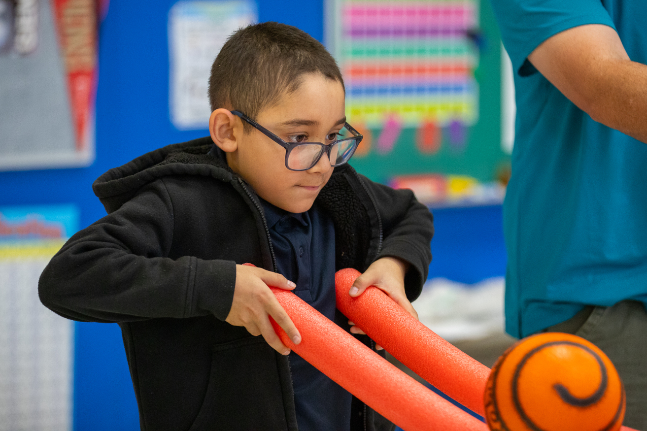 A boy with glasses plays with pool noodles and a ball in PE class.