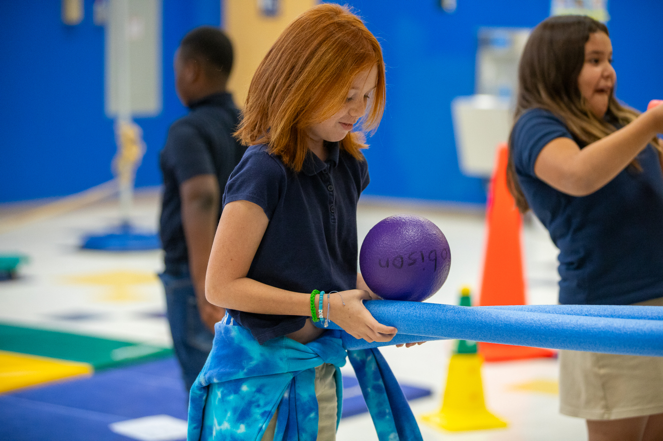 A girl with red hair plays with a ball and pool noodles in a PE STEM game.