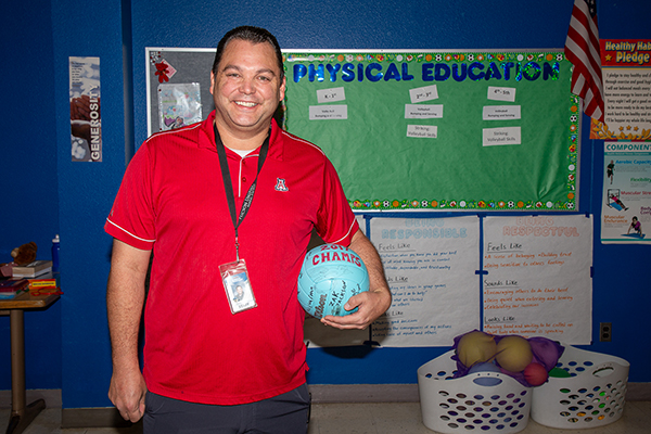 A man in a red shirt holding a light blue volleyball smiles in front of his PE board