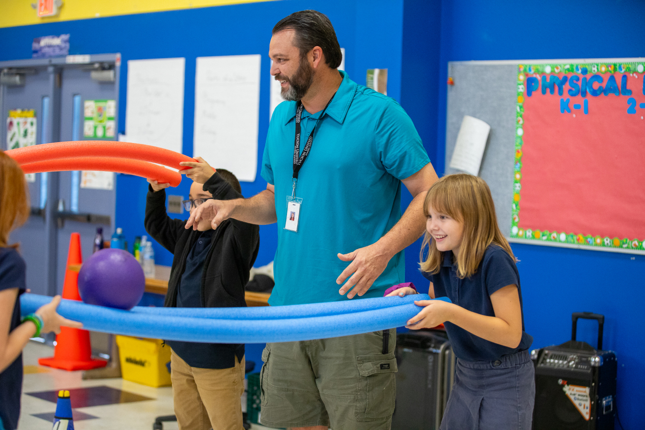 A PE teacher plays a STEM game with students using pool noodles and balls.